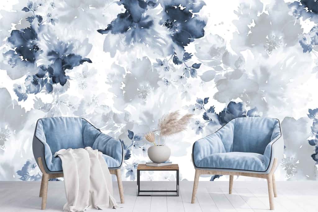 The wallpaper showcases a large floral mural in shades of blue and white, creating a calming ambiance. It features intricate patterns of blooming flowers, delicate petals, and twisting vines. The various tones of blue range from soft pastels to deep navy, interspersed with crisp white highlights that add depth and contrast. This elegant design evokes tranquility and serenity in the living room setting.