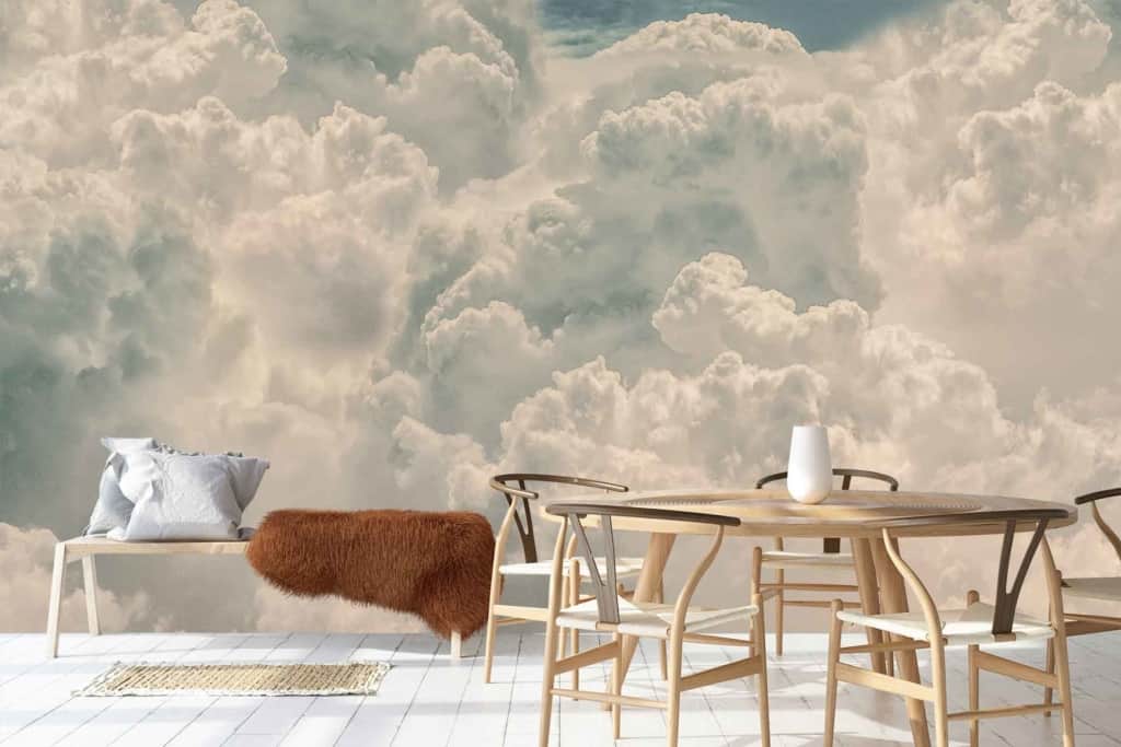 A serene room featuring a large, realistic mural wallpaper adorned with fluffy clouds set against a clear blue sky. The "Up In The Clouds" theme evokes a dreamy, open atmosphere, with soft whites and gentle blues dominating the scene.