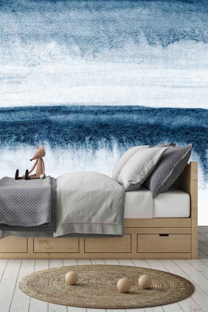 The wallpaper behind the bed is an abstract mural showcasing various shades of blue and white, creating a design reminiscent of the sea. The elements include wave-like patterns and fluid shapes, evoking a sense of movement and tranquility. Predominantly featuring hues of navy, sky blue, turquoise, and crisp white accents, the mural adds a serene backdrop to the bedroom's minimalist design.