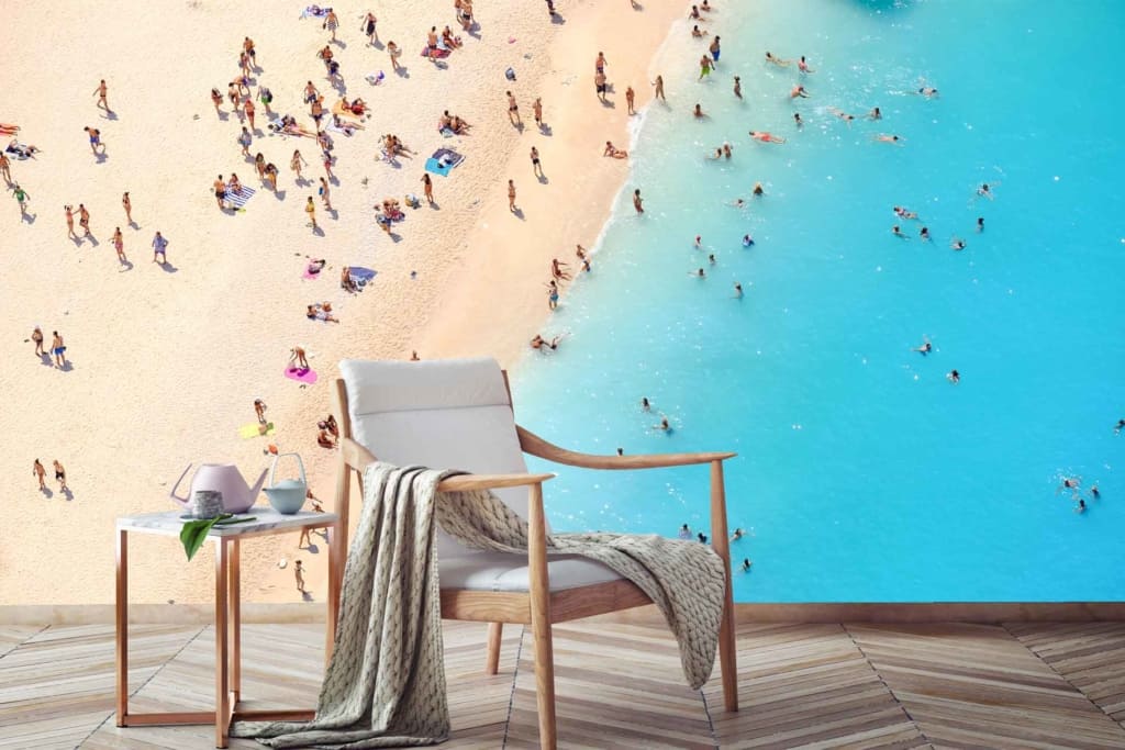 An overhead beach view wallpaper with a crowded shoreline. Dominant colors: sand beige, turquoise water, and clusters of people. Foreground features: cozy armchair with blanket, small side table with a teapot and cup on wooden floorboards.