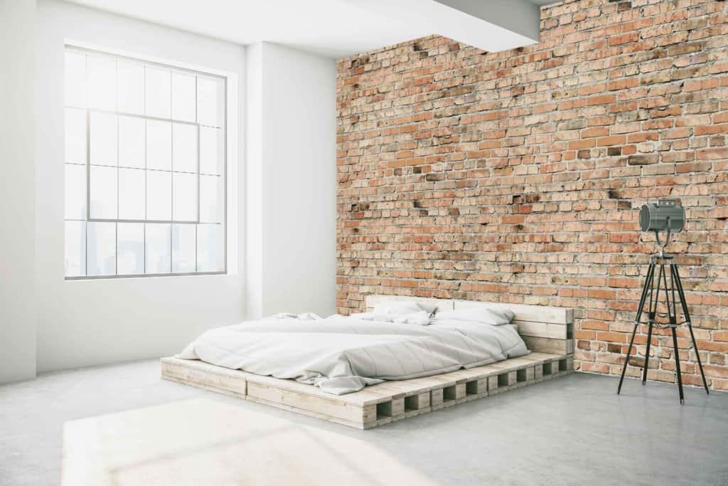 The wallpaper in the minimalist bedroom is distinguished by its large, exposed brick design, primarily featuring shades of deep red and earthy brown. Subtle variations in brick size and texture contribute to a realistic, industrial feel. The wall may also show hints of weathering or whitewashing for added depth and character, making it a striking focal point against the cool tones of the concrete floor.