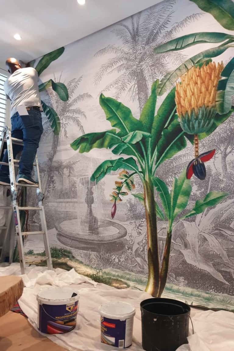 A person installs a large, tropical-themed wallpaper mural adorned with lush green banana leaves and vibrant exotic flowers. The wallpaper features rich shades of emerald, lime, and jade complemented by bursts of vivid hues such as fuchsia, sunset orange, and bright yellow from the flowers. This striking design showcases an earthy yet energetic palette perfect for any interior space.