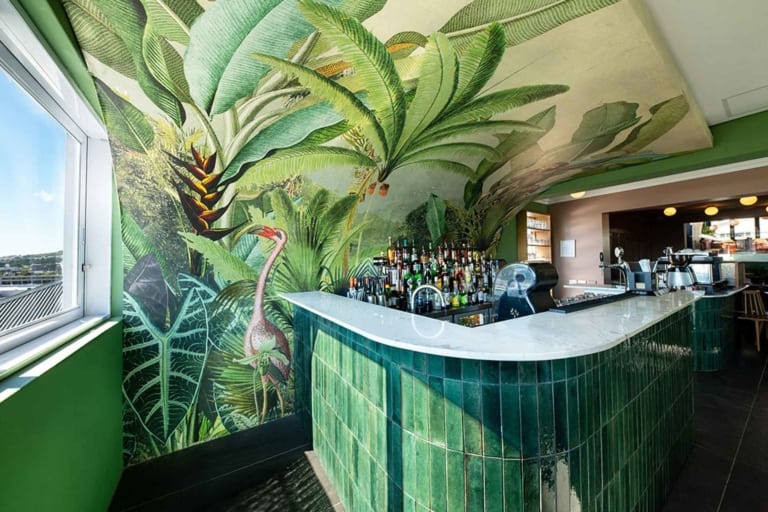 Vibrant wall mural featuring lush tropical plants in various shades of green, punctuated by colorful bird elements.