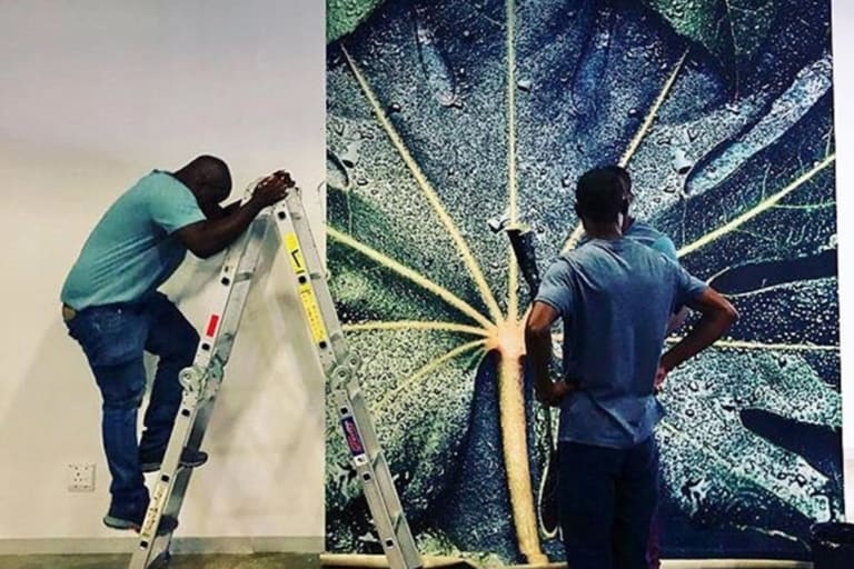 A large, detailed image of a water-covered leaf, showcasing vibrant green hues with glistening water droplets, is being hung on the wall. The wallpaper features intricate textures and shades that capture the delicate veins of the leaf and the reflective quality of the water.