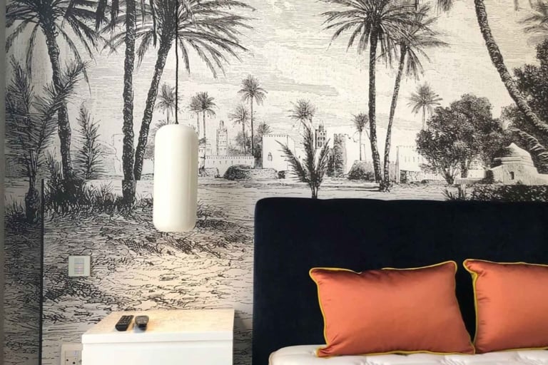 A bedroom featuring an intricate black and white wall mural of a desert scene with palm trees and buildings. The detailed wallpaper showcases elegant monochrome elements, including palm fronds, architectural structures, sandy dunes, and cloud-filled skies. This stunning visual piece creates a stark yet soothing backdrop that exudes artistic sophistication. Perfect for personal websites or portfolios seeking to highlight classic decor themes with contemporary flair.