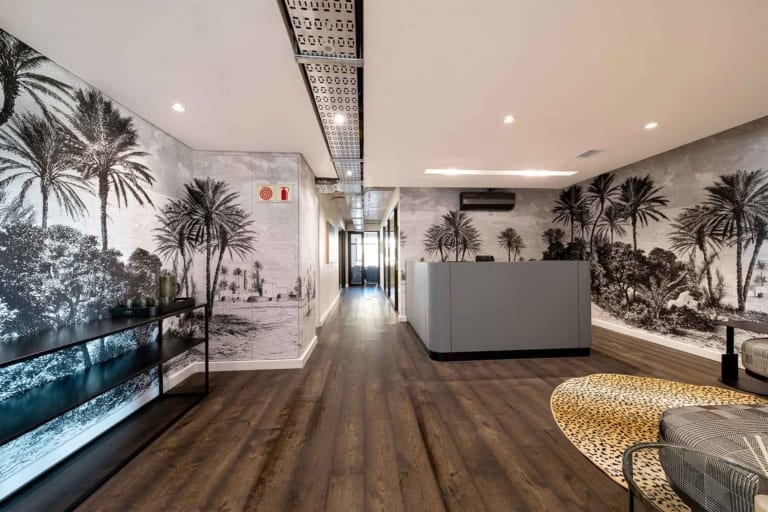 Modern office lobby features tropical wallpaper adorned with vibrant green palm leaves, lush ferns, and exotic flowers in hues of pink and yellow. The background is a subtle off-white, adding depth while letting the greenery pop. This botanical theme creates an inviting atmosphere, blending nature-inspired colors and patterns seamlessly for a fresh yet sophisticated look.