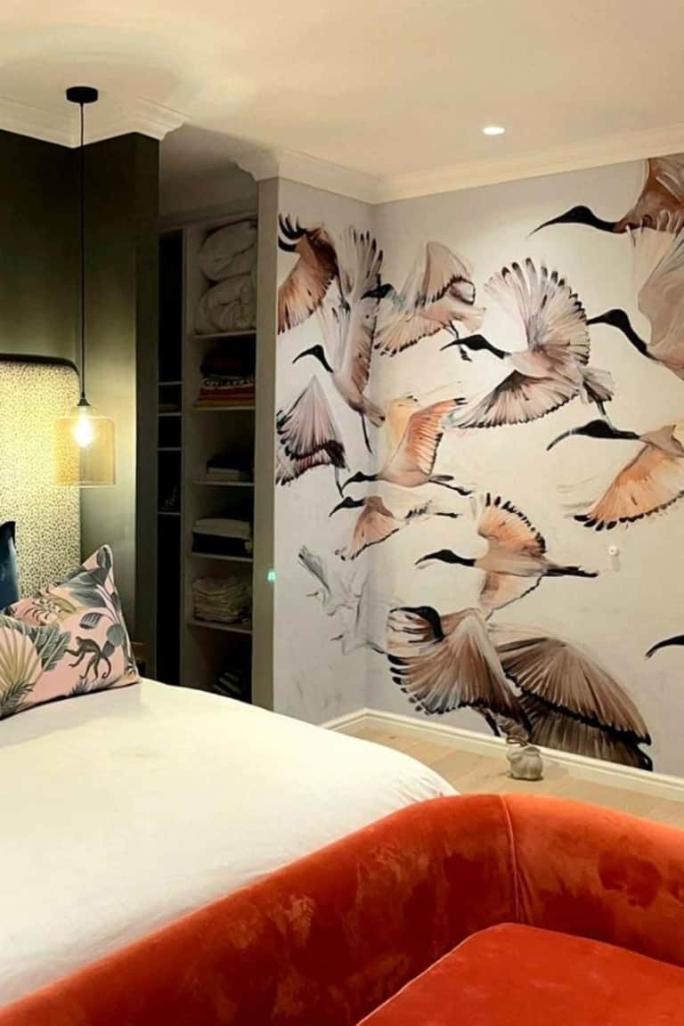A cozy bedroom showcases a mural of flying birds on the wall. The wallpaper features shades of blue skies streaked with soft white clouds, depicting birds in various stages of flight. Feather details and subtle gradients add depth, while touches of green from tree branches integrate seamlessly into the design, highlighting natural elements and tranquil colors.