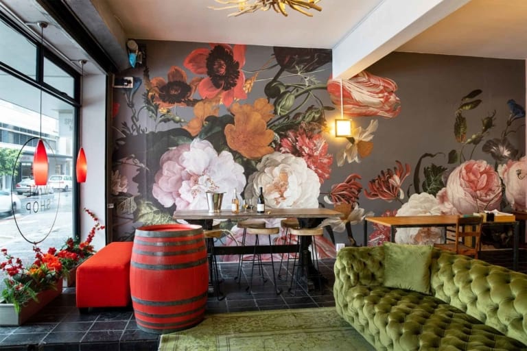 The cafe features an eye-catching floral mural with vibrant hues of pink, red, and green. The design includes intricate blossoms and lush greenery that dominate one wall. The vivid colors and detailed patterns make it a perfect backdrop for any Instagram photo.