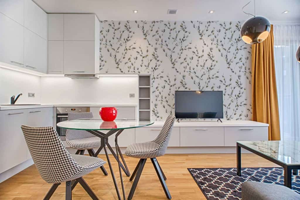 The wallpaper in the modern living and kitchen area features a floral design with swallows in mid-flight. It includes various elements such as blooms, leaves, and branches intertwined throughout. The color palette consists of soft pastels like blush pink, light blue, and mint green for the flowers and foliage. The swallows are depicted in shades of dark navy and slate gray, adding contrast against the lighter floral background. The overall theme combines nature-inspired elegance with touches of serene colors that enhance the room's ambiance.