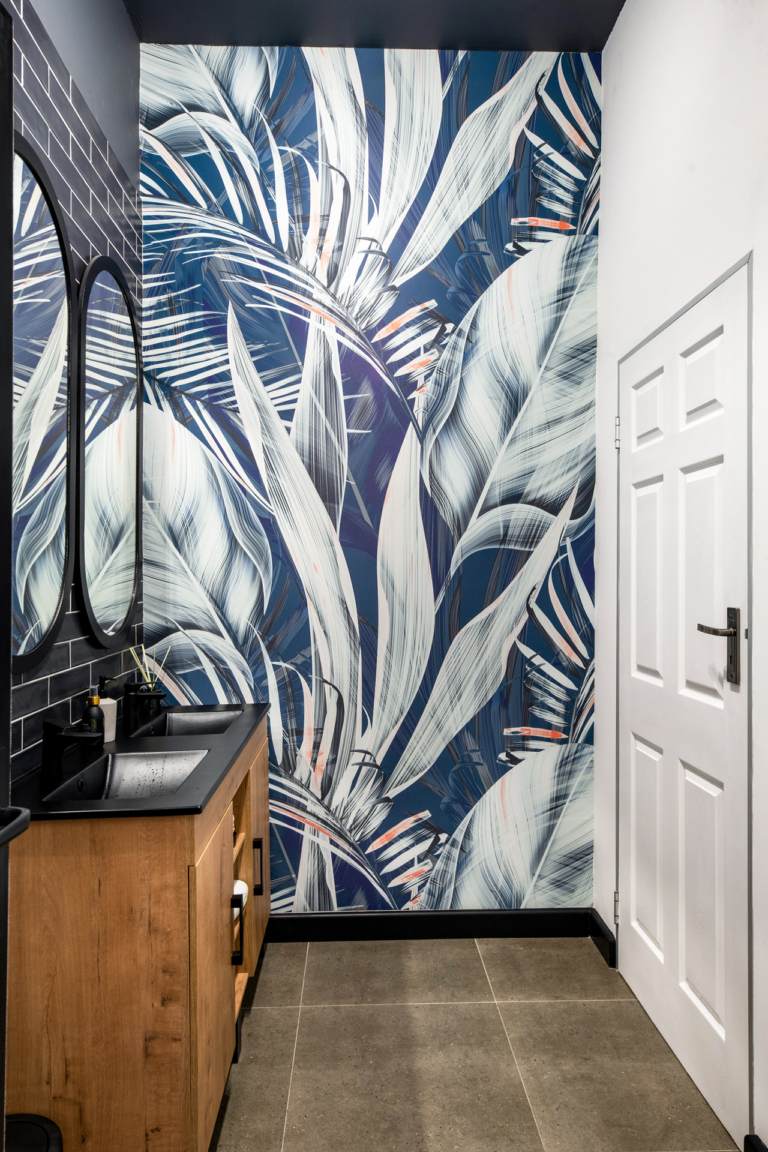 The wallpaper in this modern bathroom is adorned with vibrant, large-scale leaf patterns in striking shades of blue and white. The leaves feature intricate details, blending various hues of blue that range from deep navy to soft sky-blue. The bold design creates a stunning visual contrast against the neutral gray floor tiles, adding a lively and fresh atmosphere to the space. This eye-catching wallpaper combines elegance with nature-inspired elements, making it a standout feature in the room's decor.