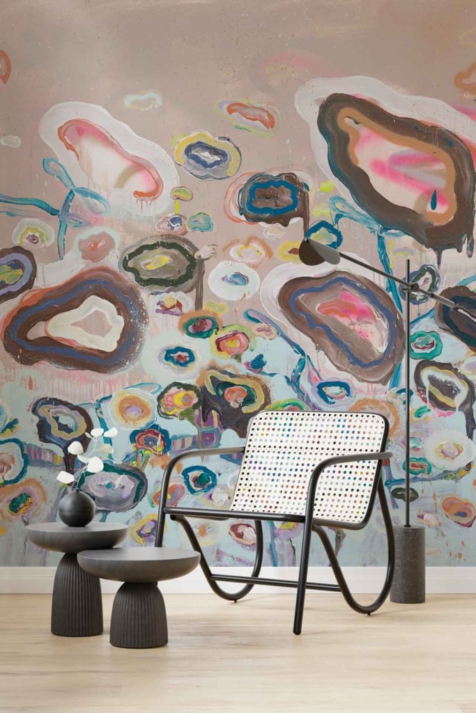 The wallpaper features a vibrant abstract mural with organic shapes, evocative of performance art. It includes bold swirls of deep blue, lively splashes of coral red, and soothing patches of mint green. The design is layered with curvilinear forms in shades of yellow and pink, interspersed with contrasting black and white accents to add depth and dynamism to the living space.