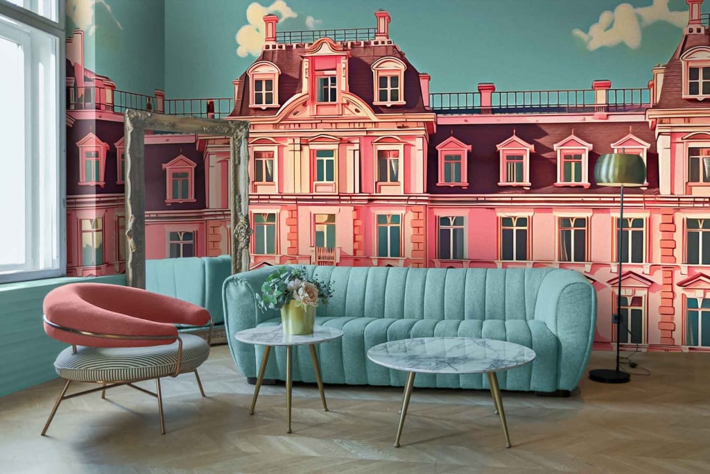 A Parisian-themed mural wallpaper featuring a pink and white building with intricate architectural details. The scene includes delicate balconies with wrought-iron railings, charming window shutters, and pastel accents that evoke a vintage European charm. Colors predominantly include pink, white, black ironwork, and hints of soft pastels creating an elegant yet whimsical backdrop reminiscent of Wes Anderson's signature style.