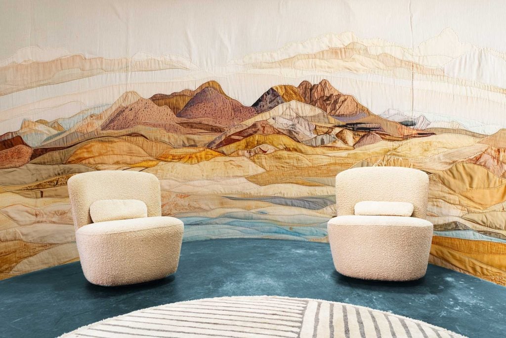 The wallpaper features a tapestry mural of mountains with warm hues, such as orange, red, and gold. It highlights natural and earthy elements, creating an artistic backdrop.