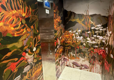 The elevator's interior is wrapped in a vibrant floral and plant-themed wallpaper. The design showcases bold, colorful abstract depictions of various flowers like lilies, roses, and daisies intertwined with lush greenery. Shades of deep magenta, rich emerald green, sunny yellow, soft lavender, and vibrant coral dominate the pattern. Intricate vines and leaves weave through the scene with accents in bright turquoise and gentle creams to add contrast to the lively floral motifs. This striking display creates an enchanting garden ambiance within the confined space.