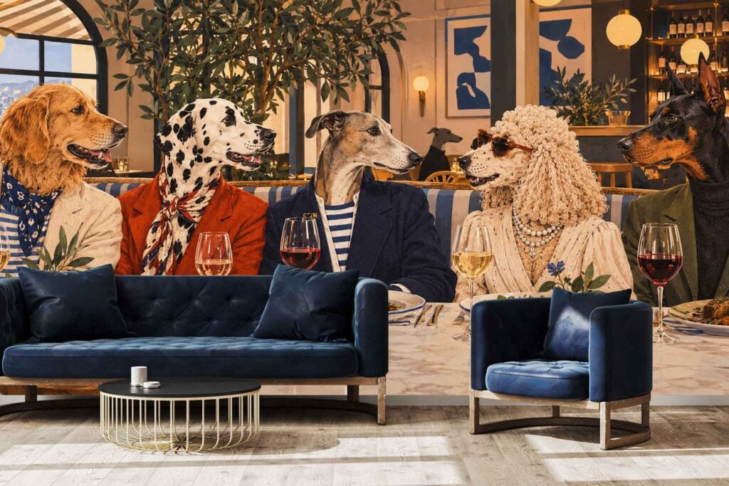 Wallpaper showcases five fashionable dogs at a restaurant table, dressed in jackets and accessories with wine glasses and plates. The mural features elegant decor elements—lamps, lush green plants, and a refined palette of blues from the sofa and chair—with accents of gold and velvet for a luxe atmosphere.